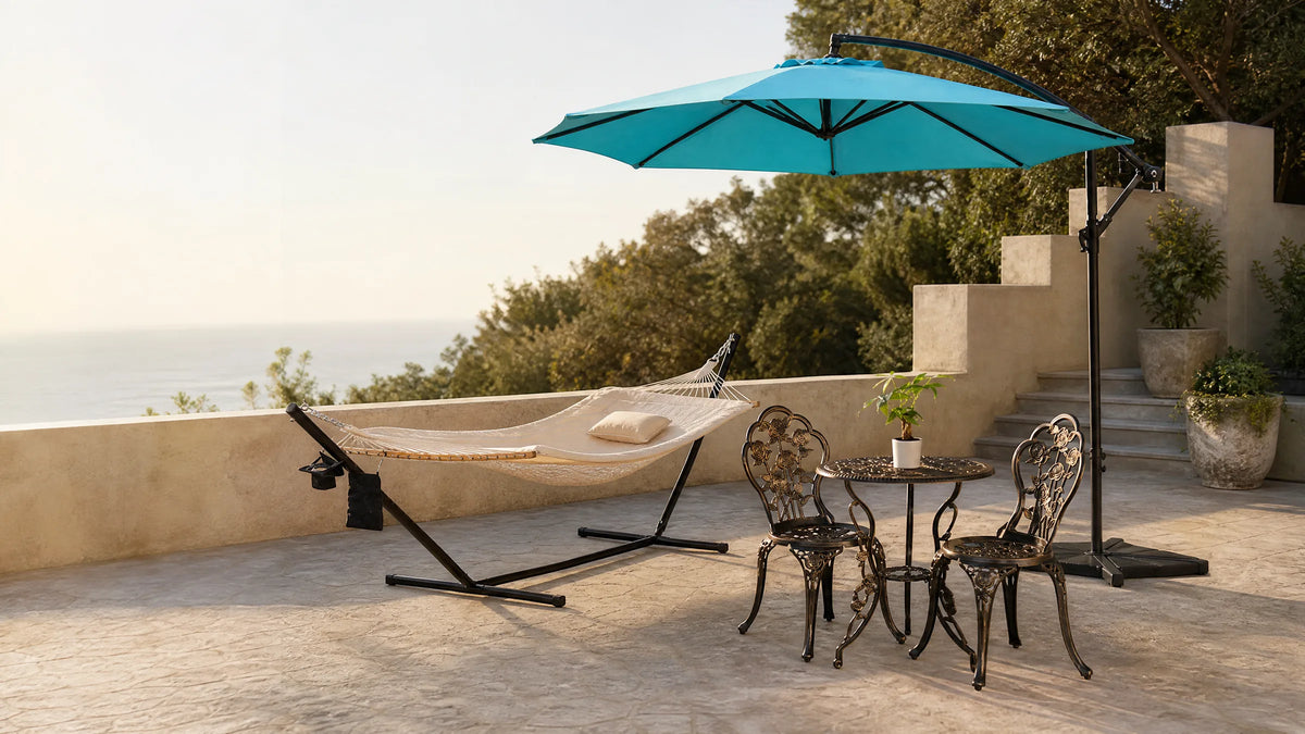 Sunlit stone patio with a hammock on a stand, a cast aluminium bistro set and an open cantilever umbrella overlooking the sea
